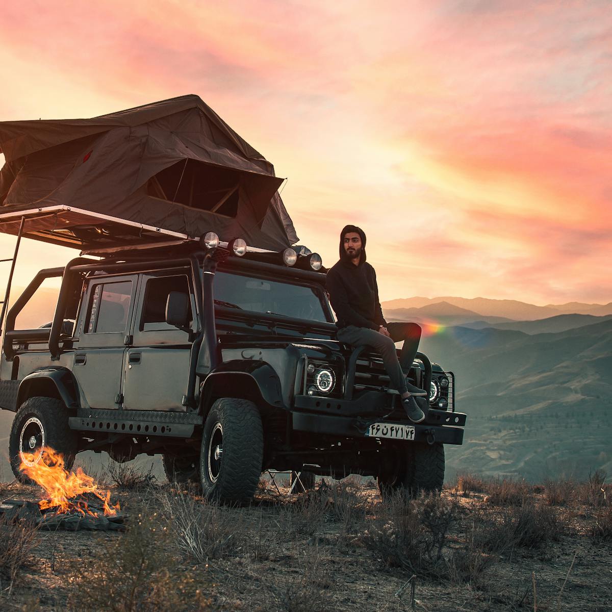 Man sitting on SUV with rooftop tent by a campfire, enjoying the sunset over mountains.